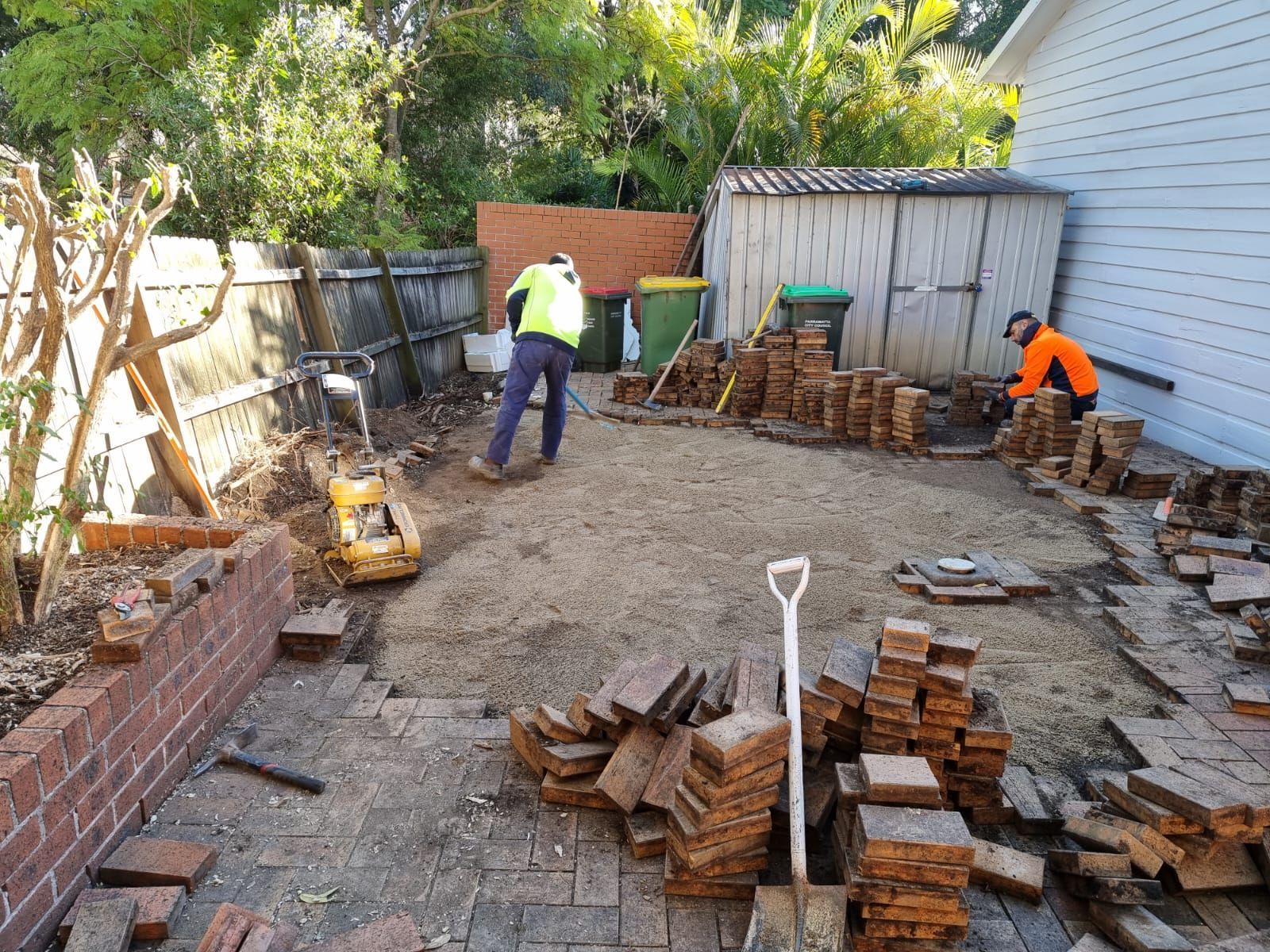 Two men are working on a brick patio in a backyard.