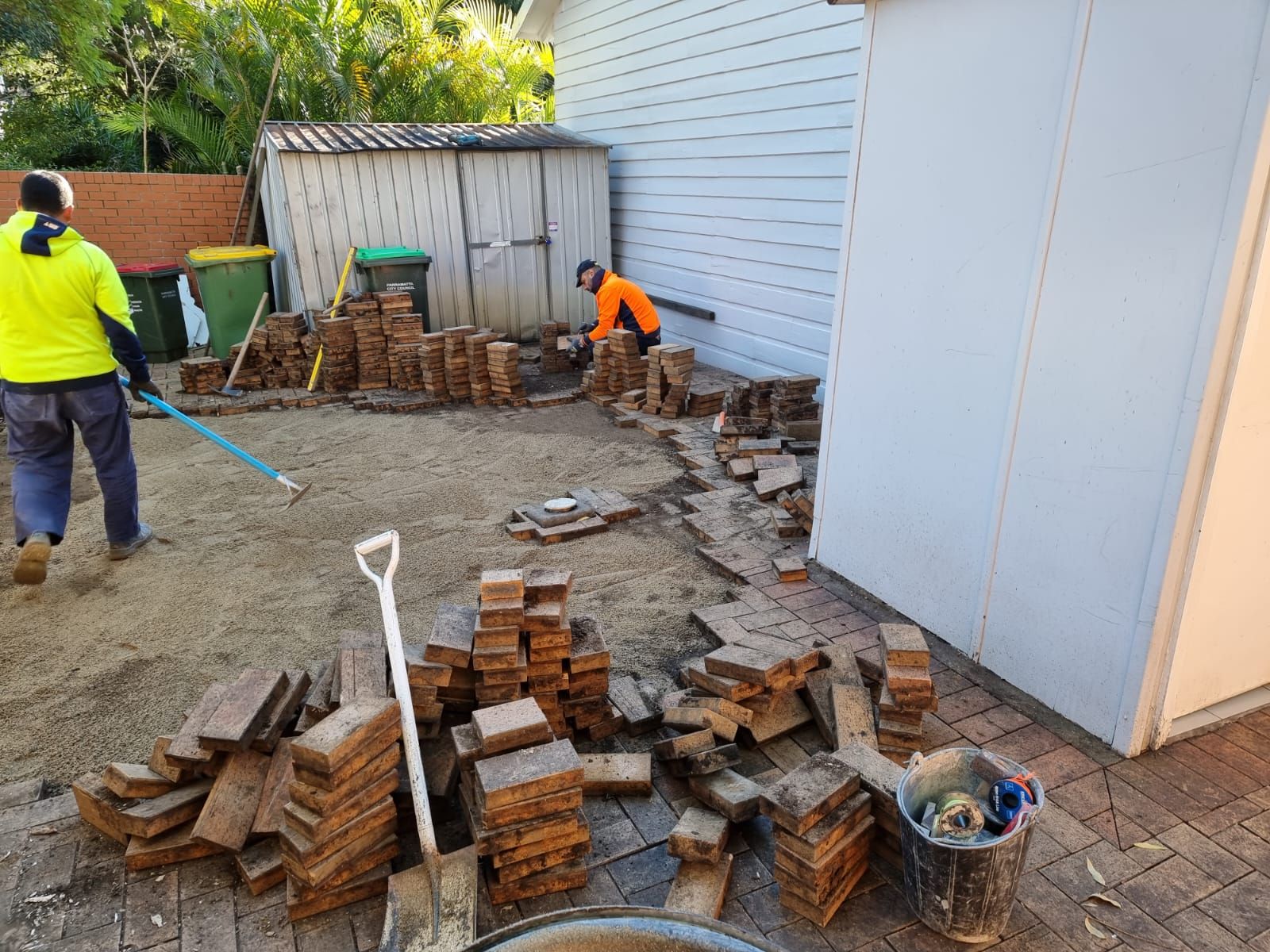 Two men are working on a brick walkway in a backyard.