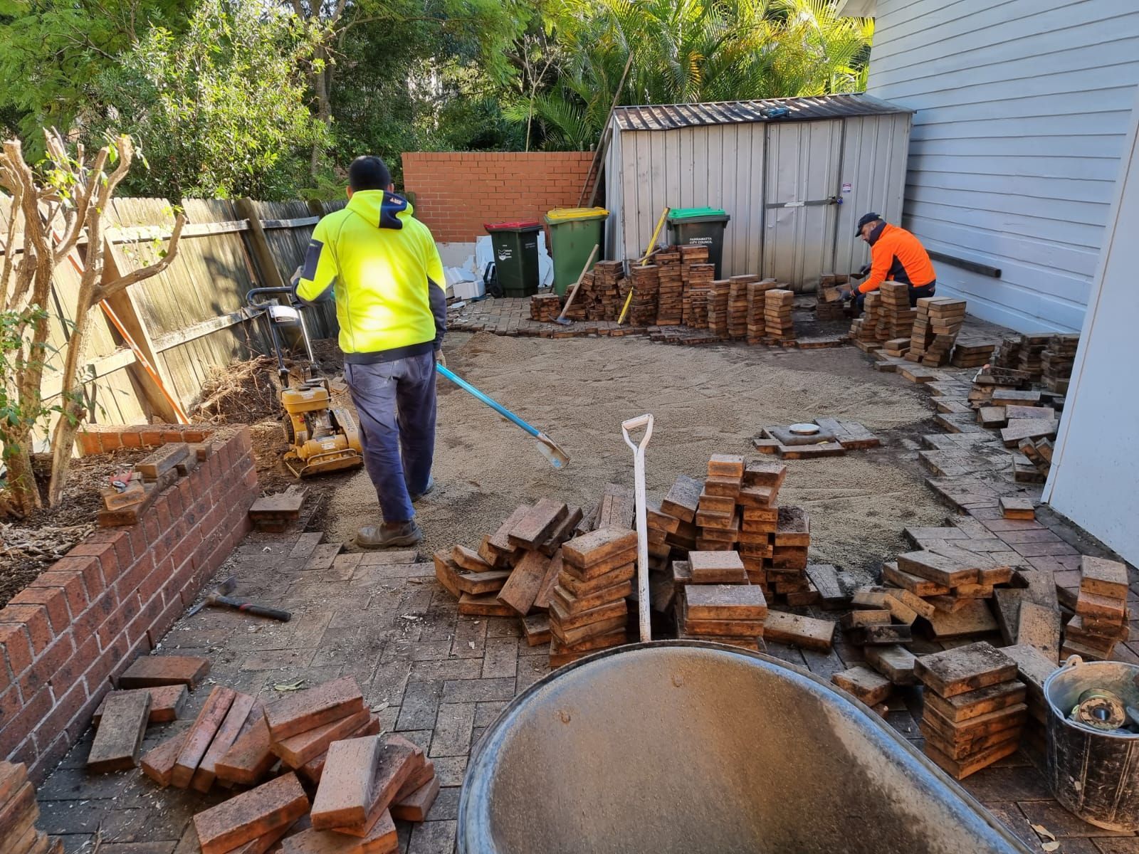 Two men are working in a backyard with bricks and a wheelbarrow.
