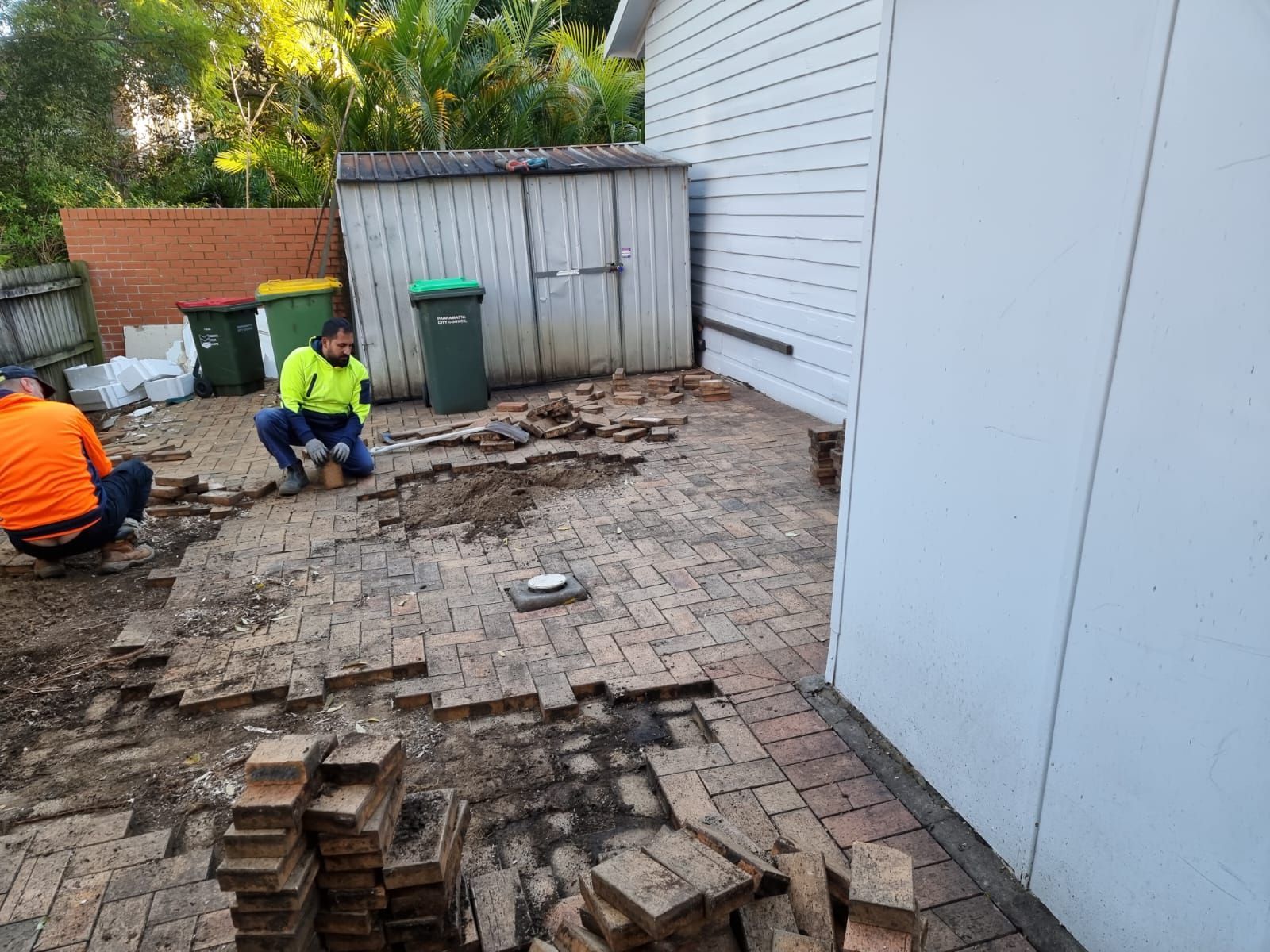 Two men are working on a brick patio in front of a house.