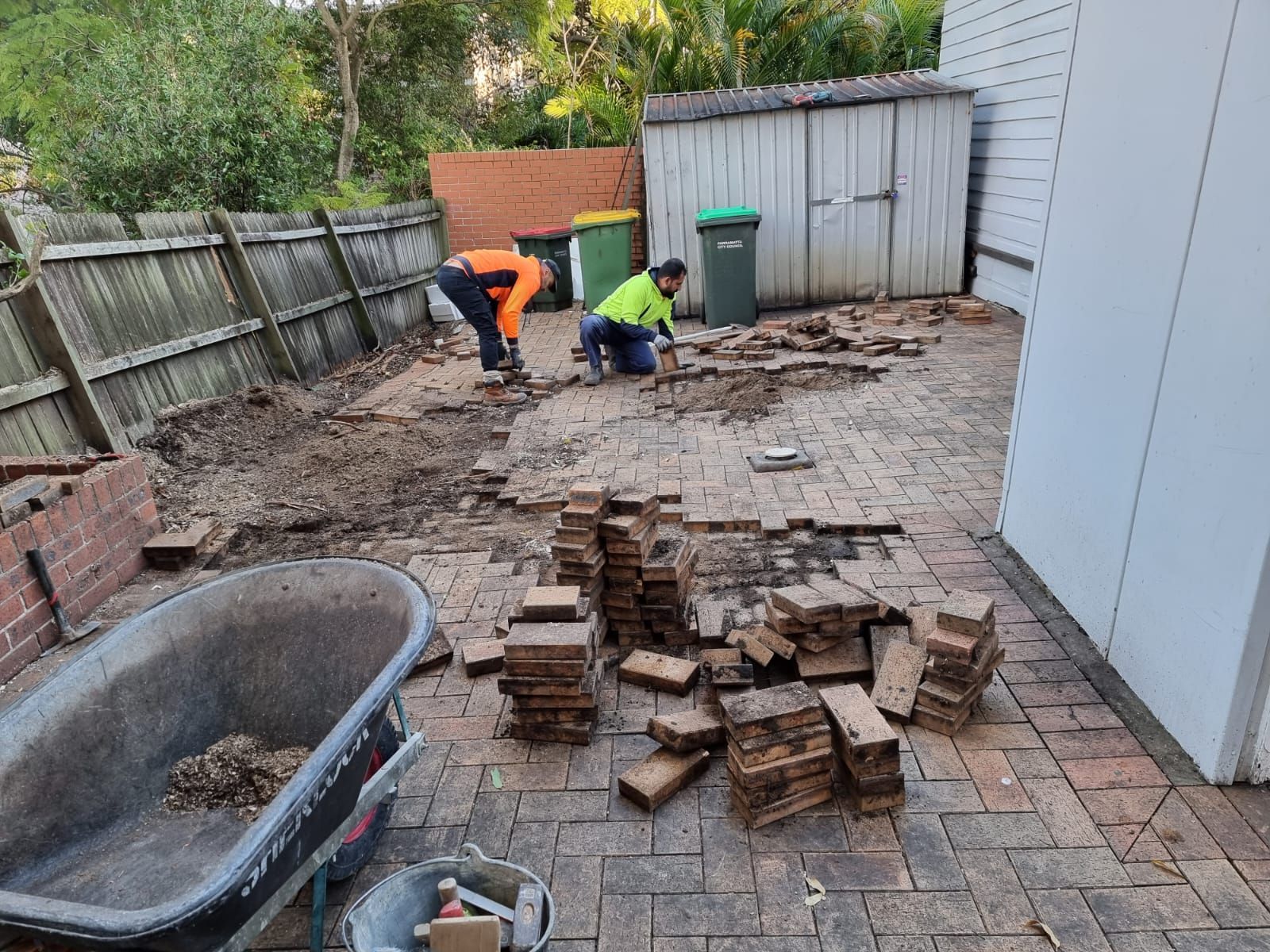 Two men are working on a patio with bricks and a wheelbarrow.