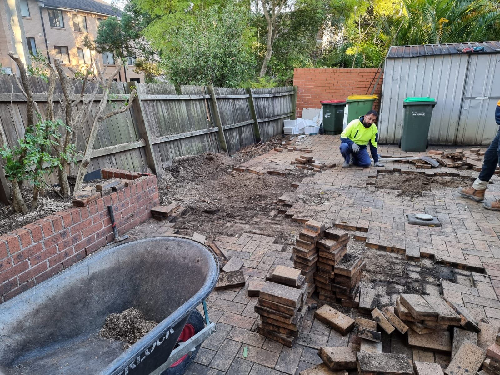 A man is kneeling on a brick patio next to a wheelbarrow.