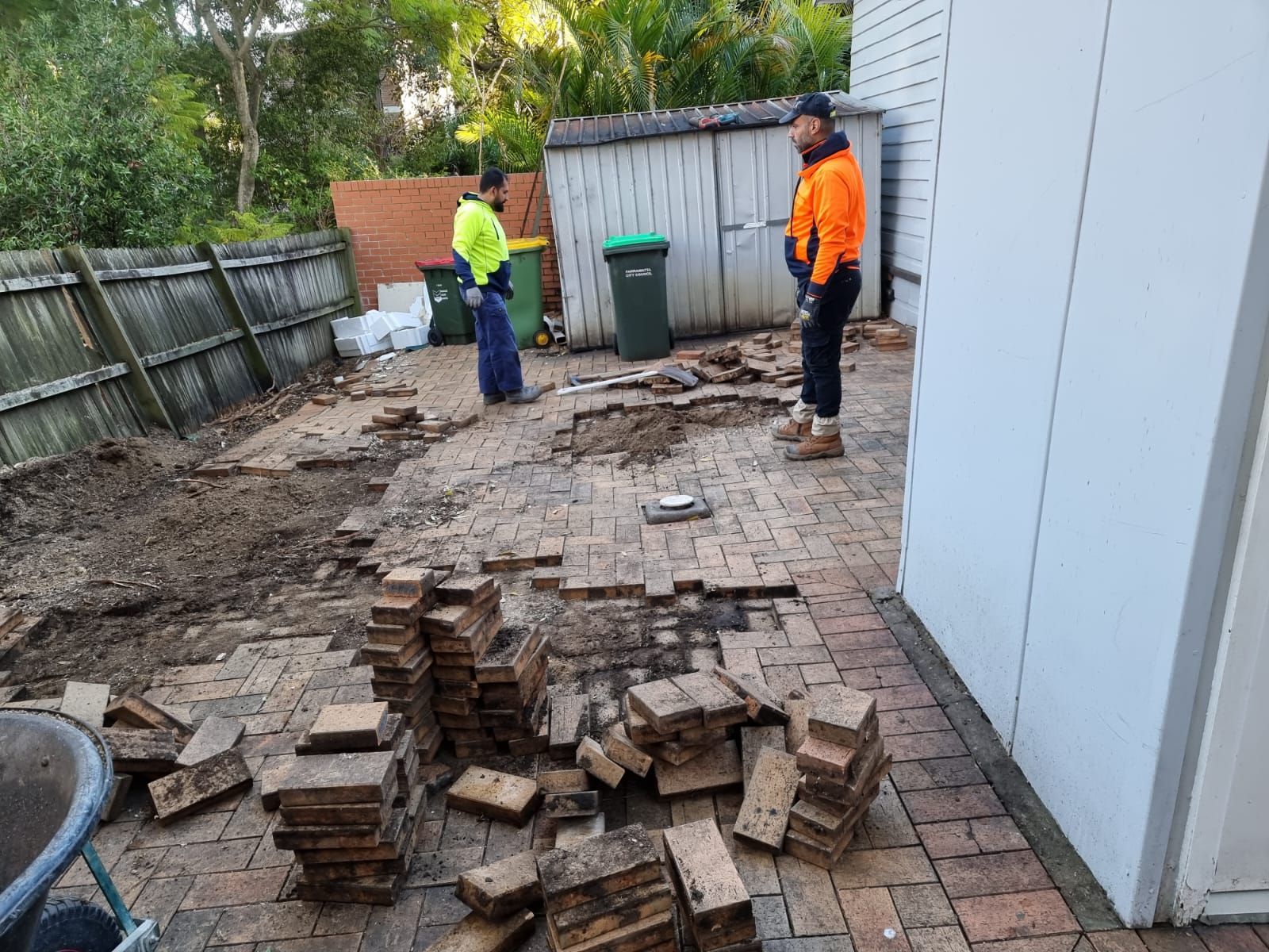Two men are standing next to a pile of bricks in a backyard.