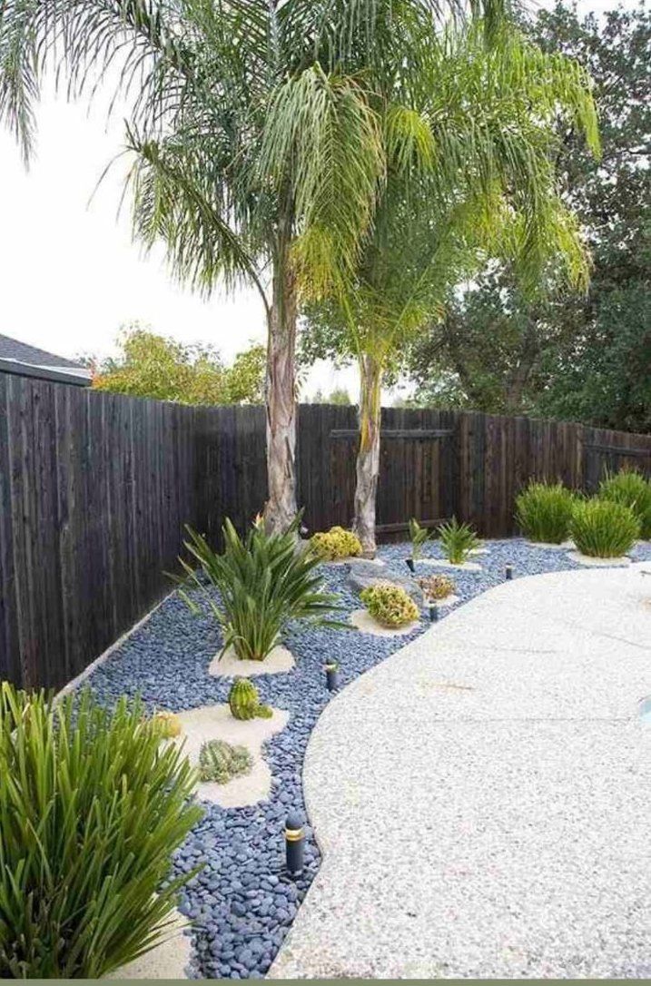 A backyard with a fence , palm trees , gravel and plants.