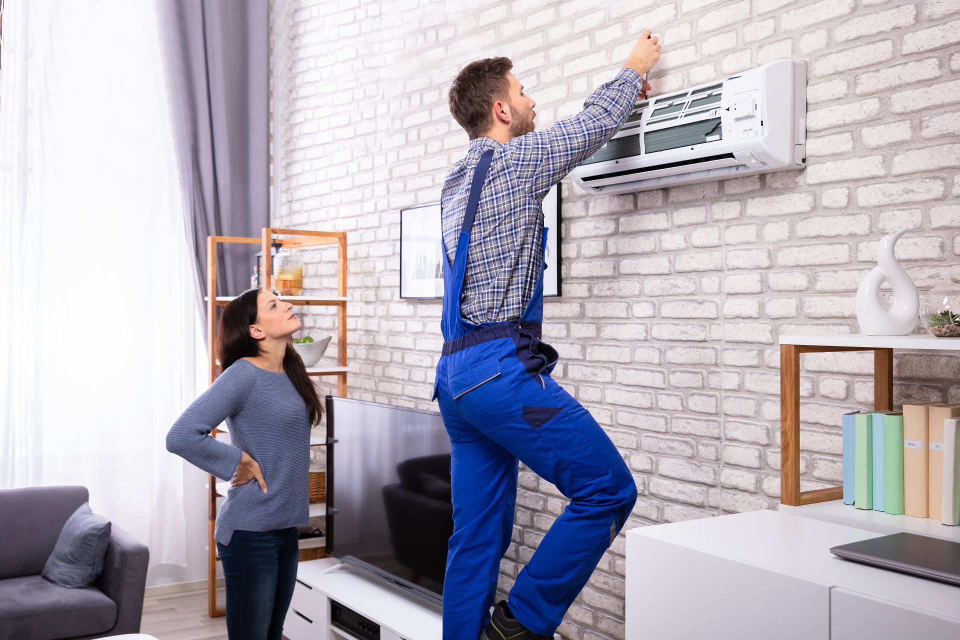Young Woman Looking At Male Technician Repairing Air Conditioner Mounted On Brick Wall