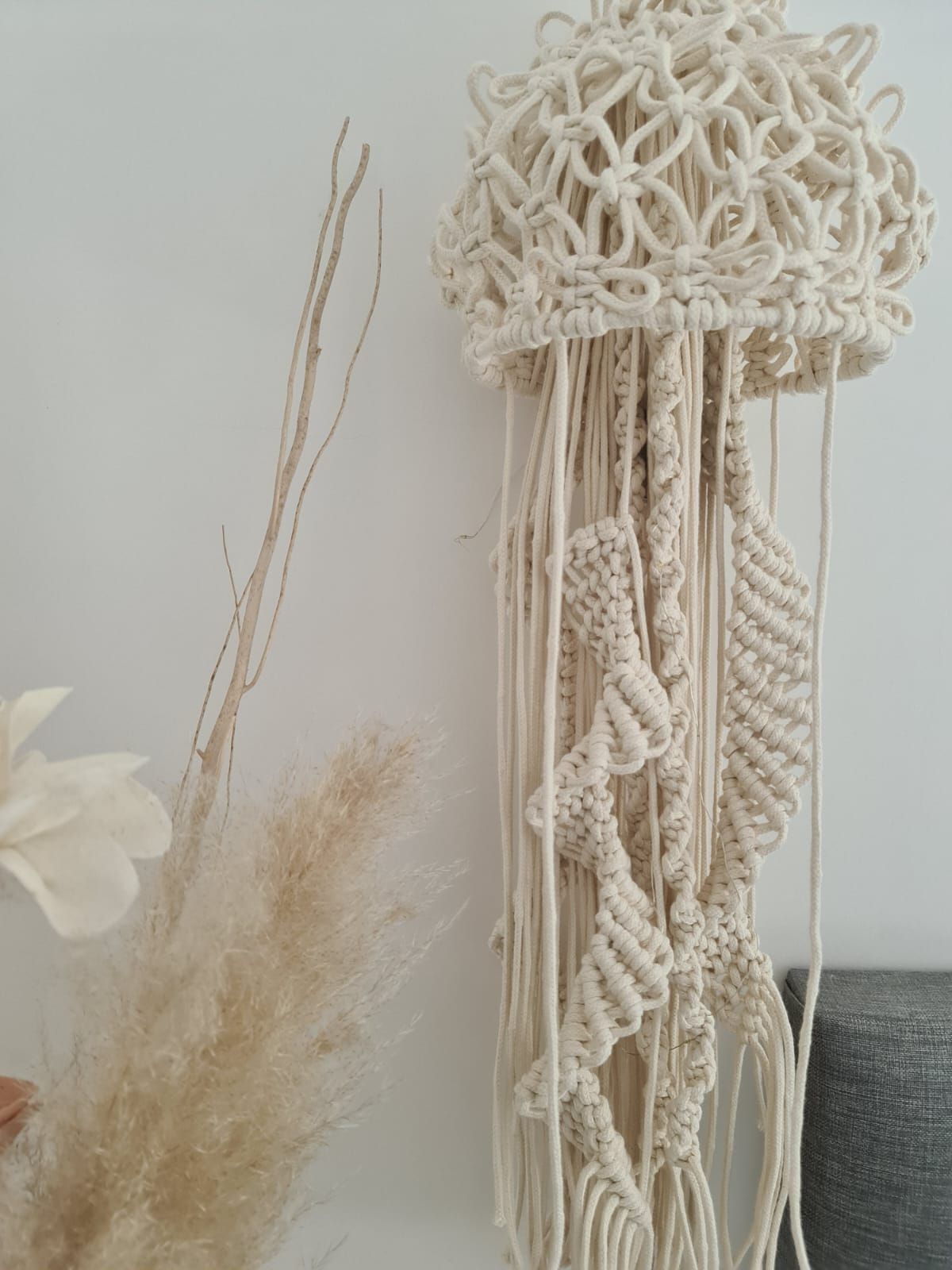 Medusa colgante de pared en macramé de color crema con tapa texturizada y hebras colgantes, junto a plantas secas.