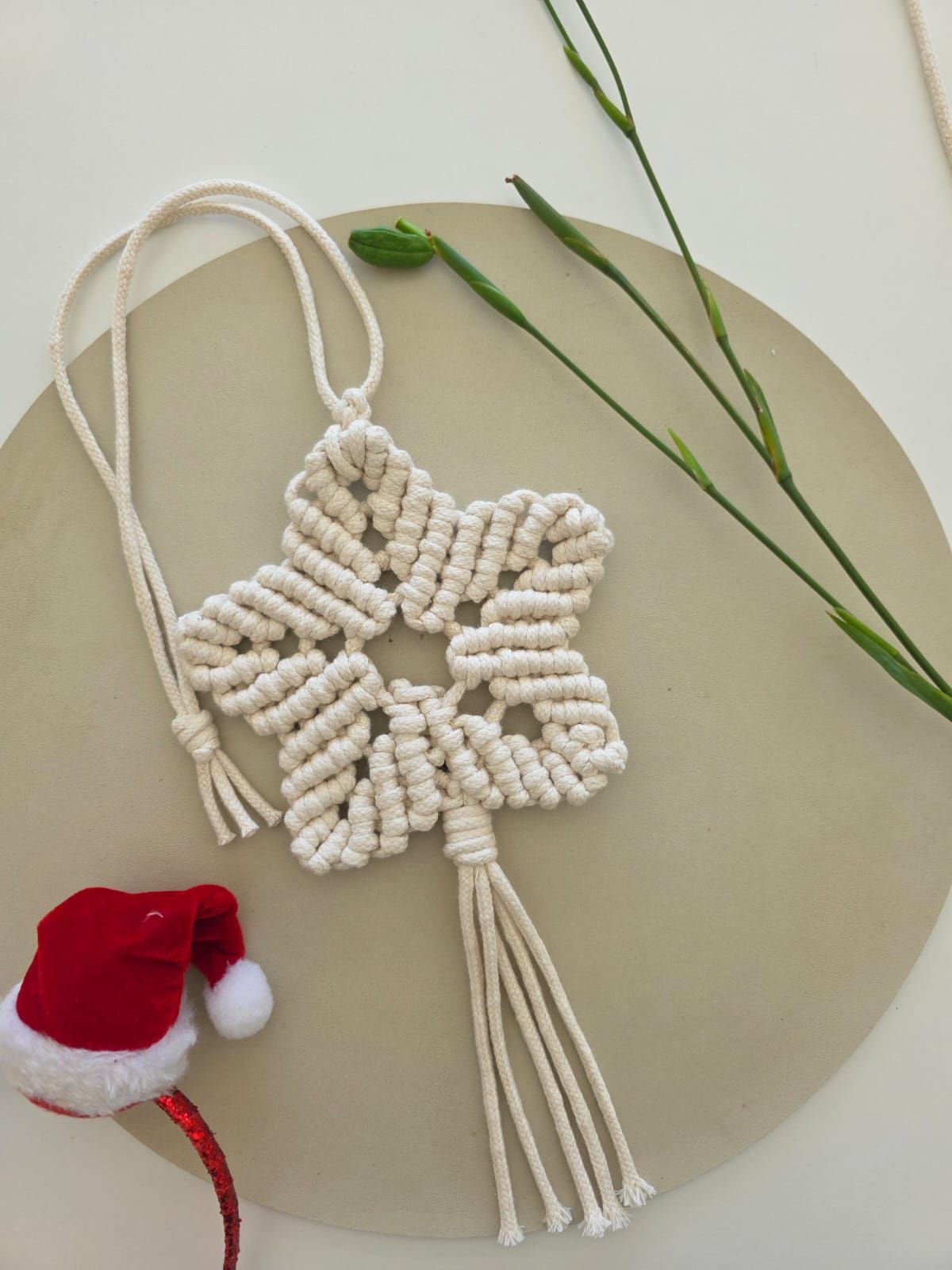Adorno de estrella de macramé con cordón color crema, colgado sobre un plato beige, al lado de un gorro de Papá Noel.