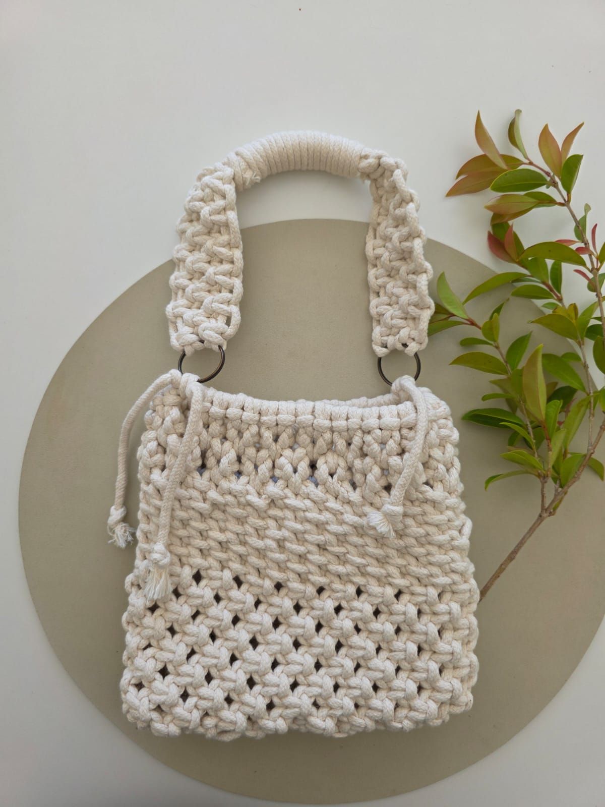 Bolso de macramé de color crema con asas, colocado sobre una superficie redonda, junto a ramas de plantas verdes.