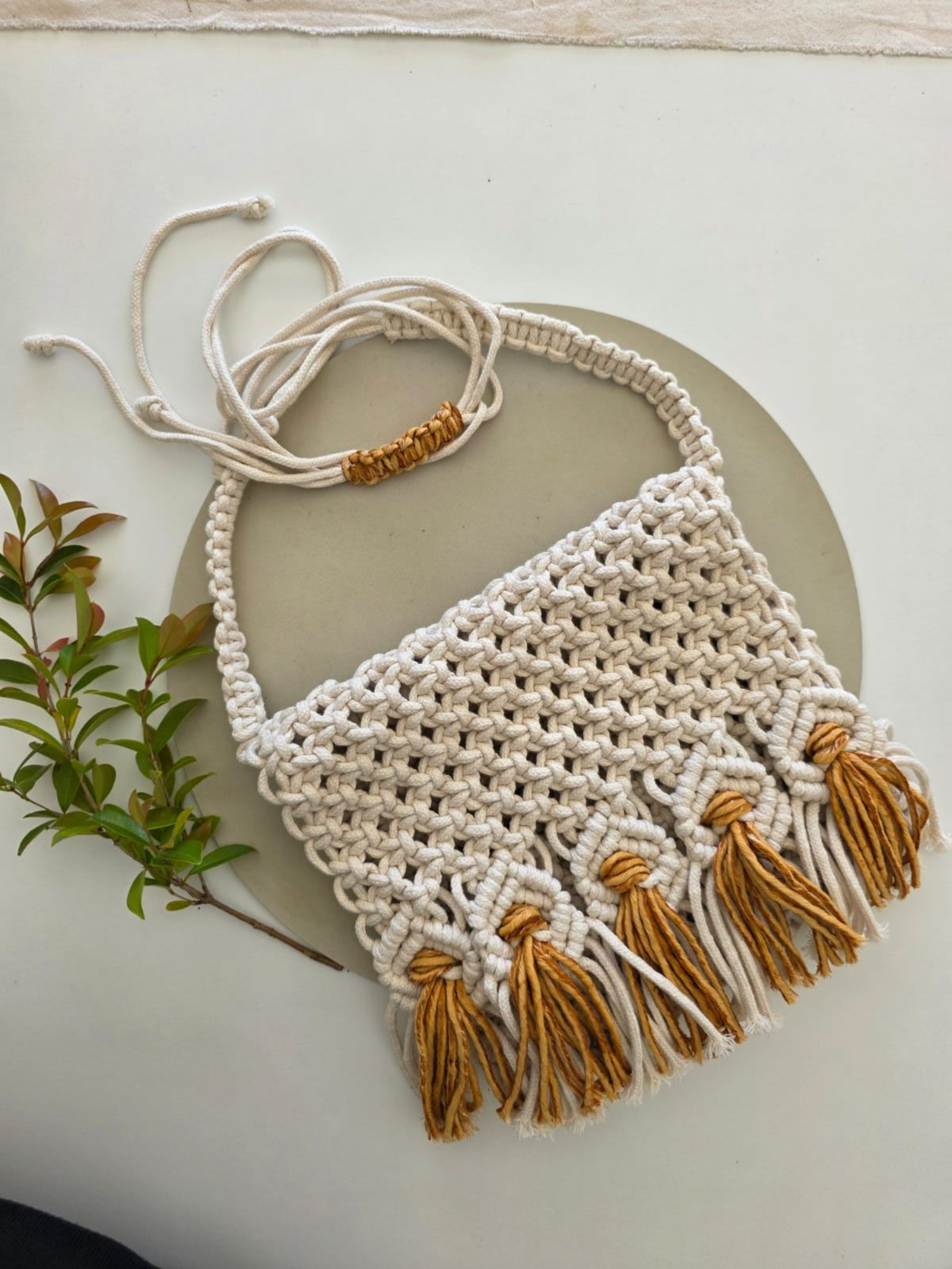 Monedero de macramé con flecos de color amarillo mostaza, cordón crema y marrón, sobre fondo neutro con verdes.