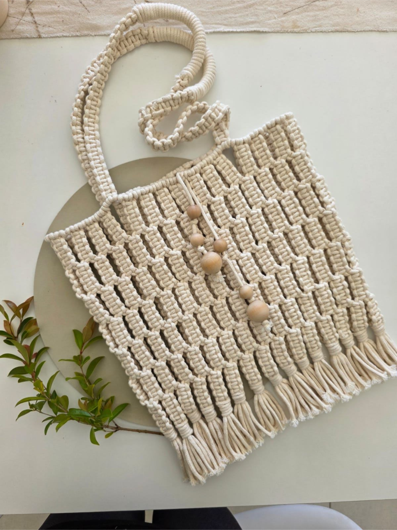 Bolso tote de macramé con flecos, color beige, con cuentas de madera y ramita de hojas sobre superficie blanca.
