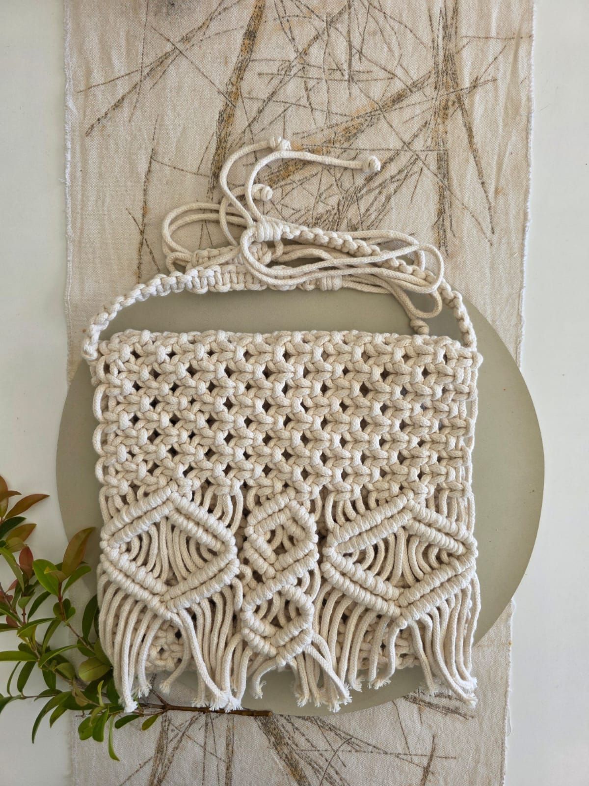 Bolso bandolera de macramé en color natural, con dibujos geométricos y flecos sobre fondo neutro.