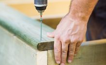 a builders hand as he is holding down a piece of wood to drill