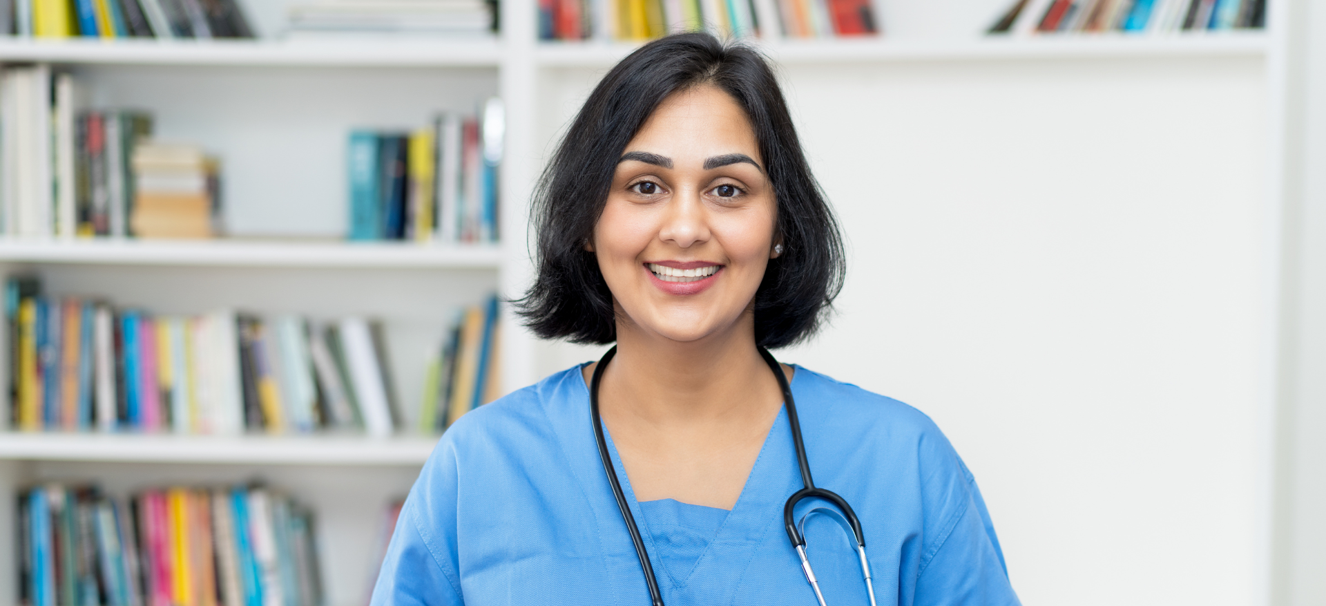 A female doctor with a stethoscope around her neck is smiling in front of a bookshelf.