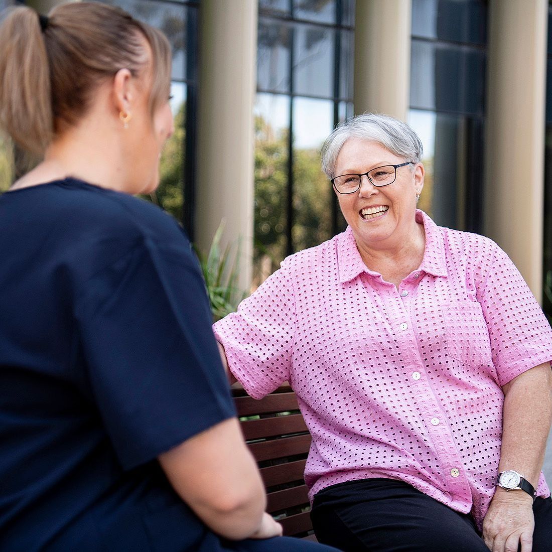 Two women are sitting on a bench talking to each other . one of the women is wearing glasses and a pink shirt.