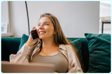 Woman smiling while talking on a phone, sitting on a green couch in a brightly lit room.