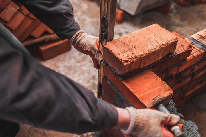 Person laying bricks, using a level, outdoors. Red bricks, grey mortar, gloved hands.