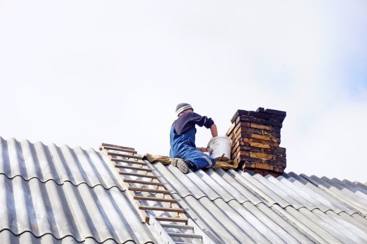 Person on a roof near a chimney, working with a bucket. Gray roof, blue sky.