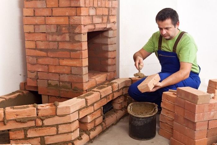 Man building brick fireplace, wearing blue overalls, placing brick with mortar. Interior setting.