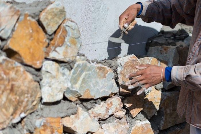 Person using a trowel to place a stone in a wall, mortar visible, outdoors.