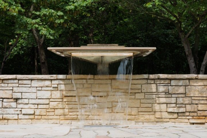 Water cascading from a square concrete structure into a stone wall fountain. Trees in the background.
