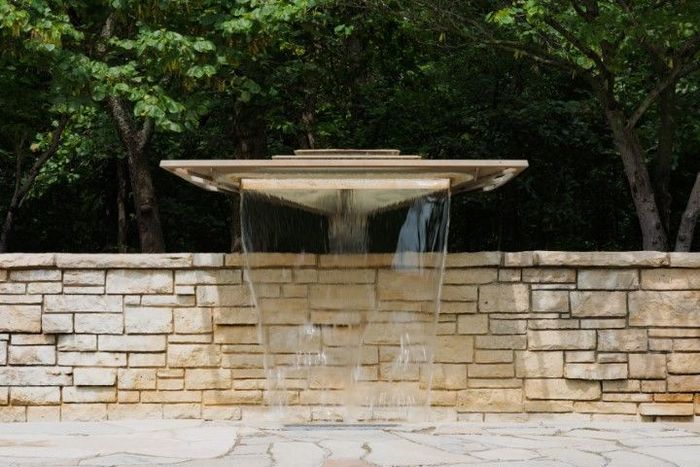 Water cascading from a square concrete structure into a stone wall fountain. Trees in the background.