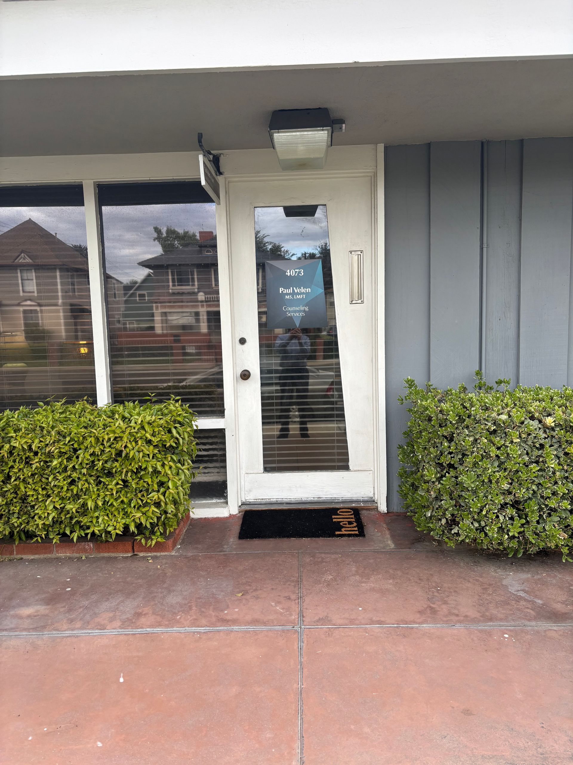 Entrance to a business with glass door, windows, and bushes. A person is reflected in the door.
