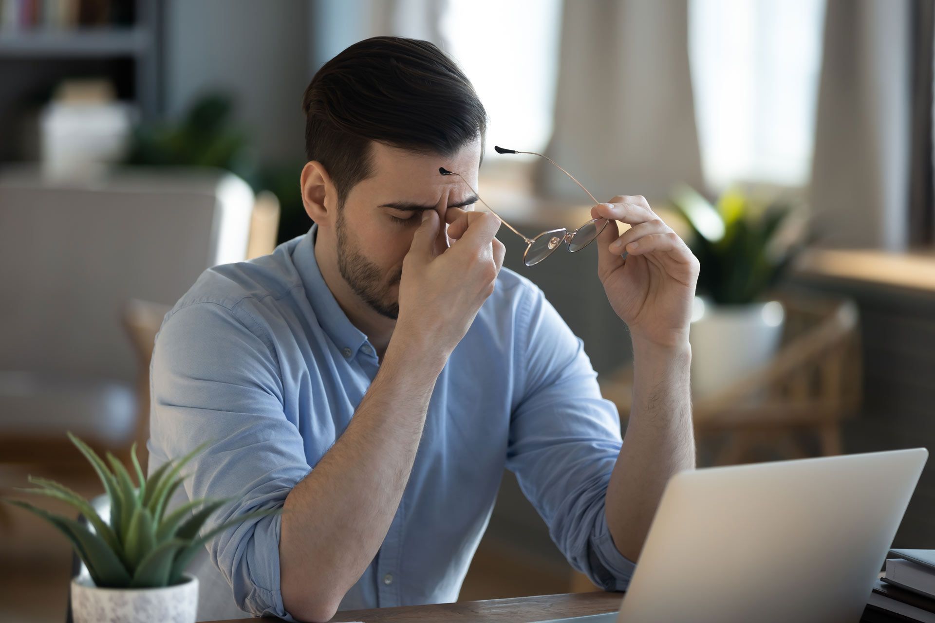 Man in blue shirt, holding glasses, rubs eyes, appearing stressed while sitting at a laptop.
