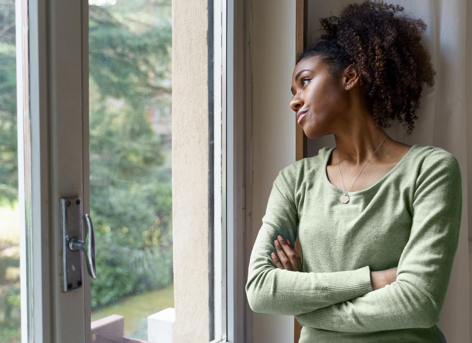 Woman with arms crossed, looking out a window at greenery.