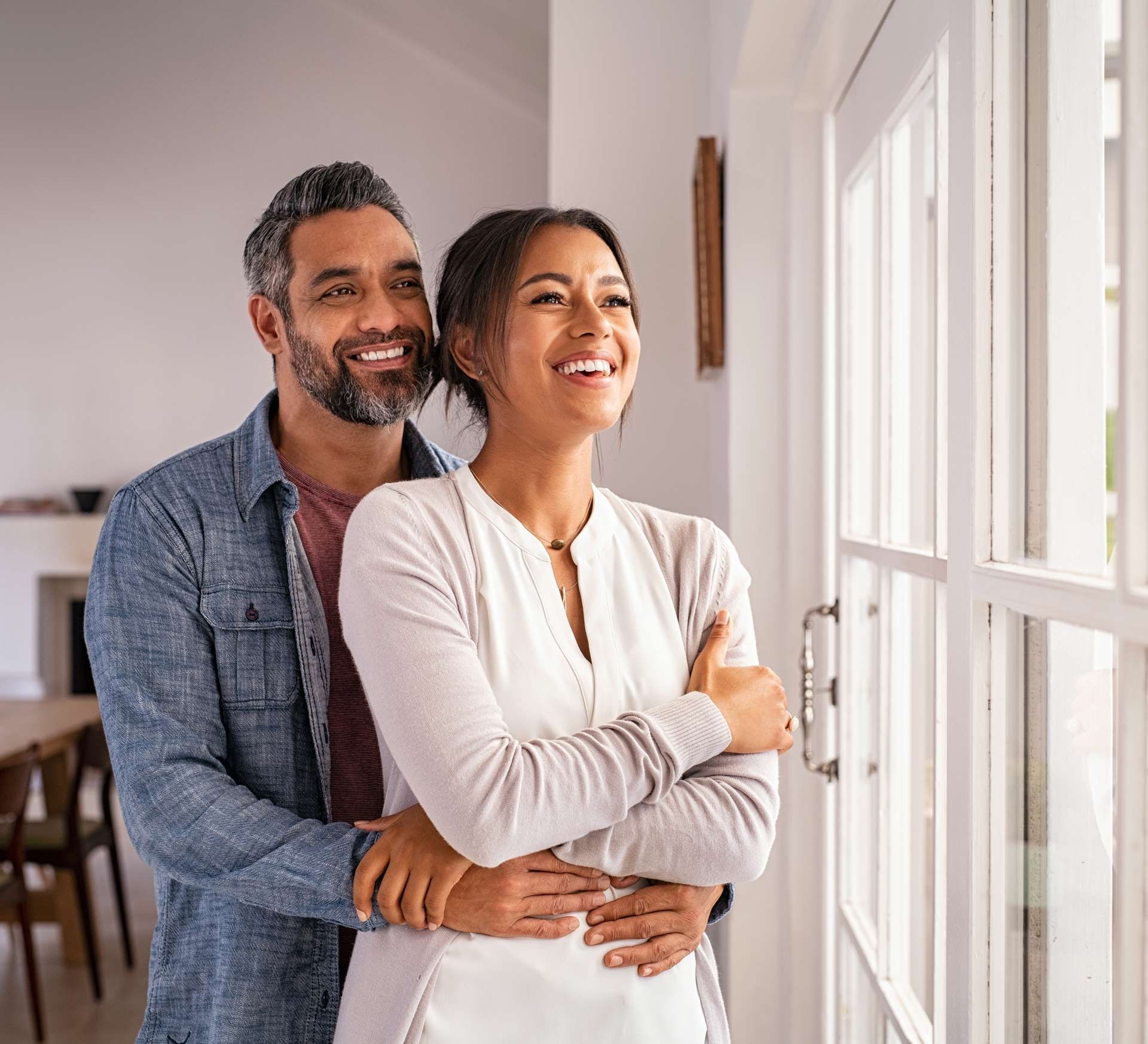 Man embraces woman by a window, both smiling and looking outward.