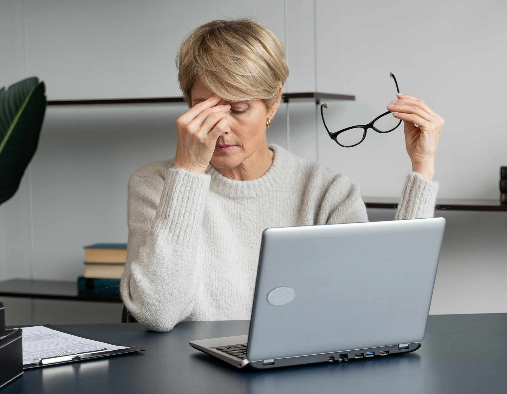 Woman at a desk, holding glasses, rubbing eyes, looking tired at a laptop.