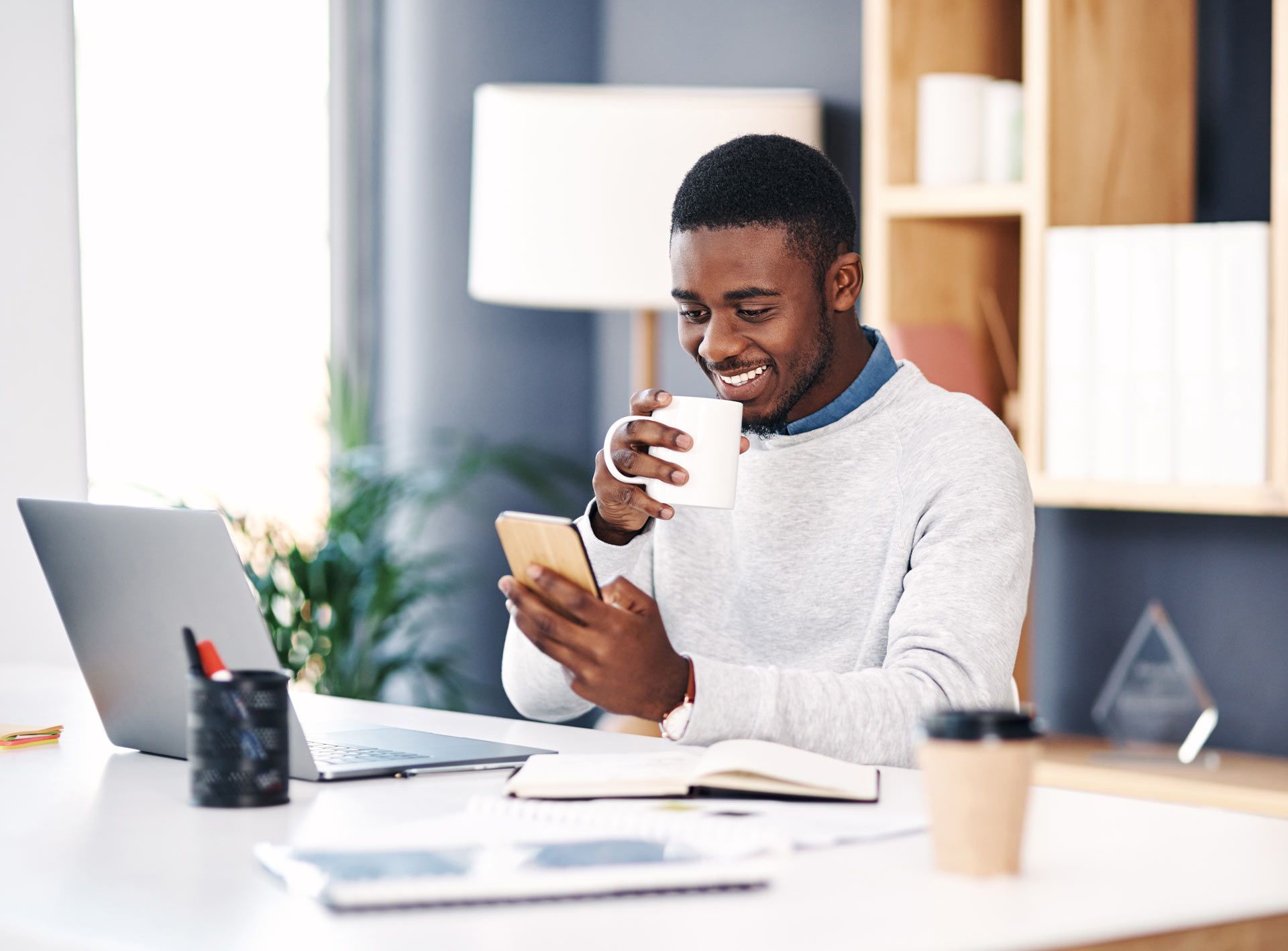 Man smiles while looking at a phone, holding a mug and sitting at a desk with a laptop.
