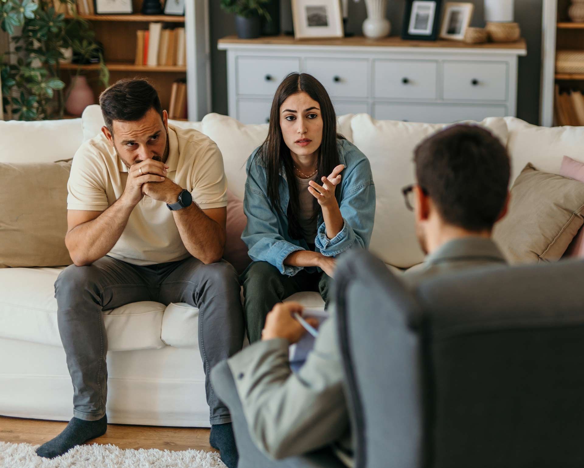 Couple on a sofa in counseling session, facing therapist. Woman gestures, man looks concerned.