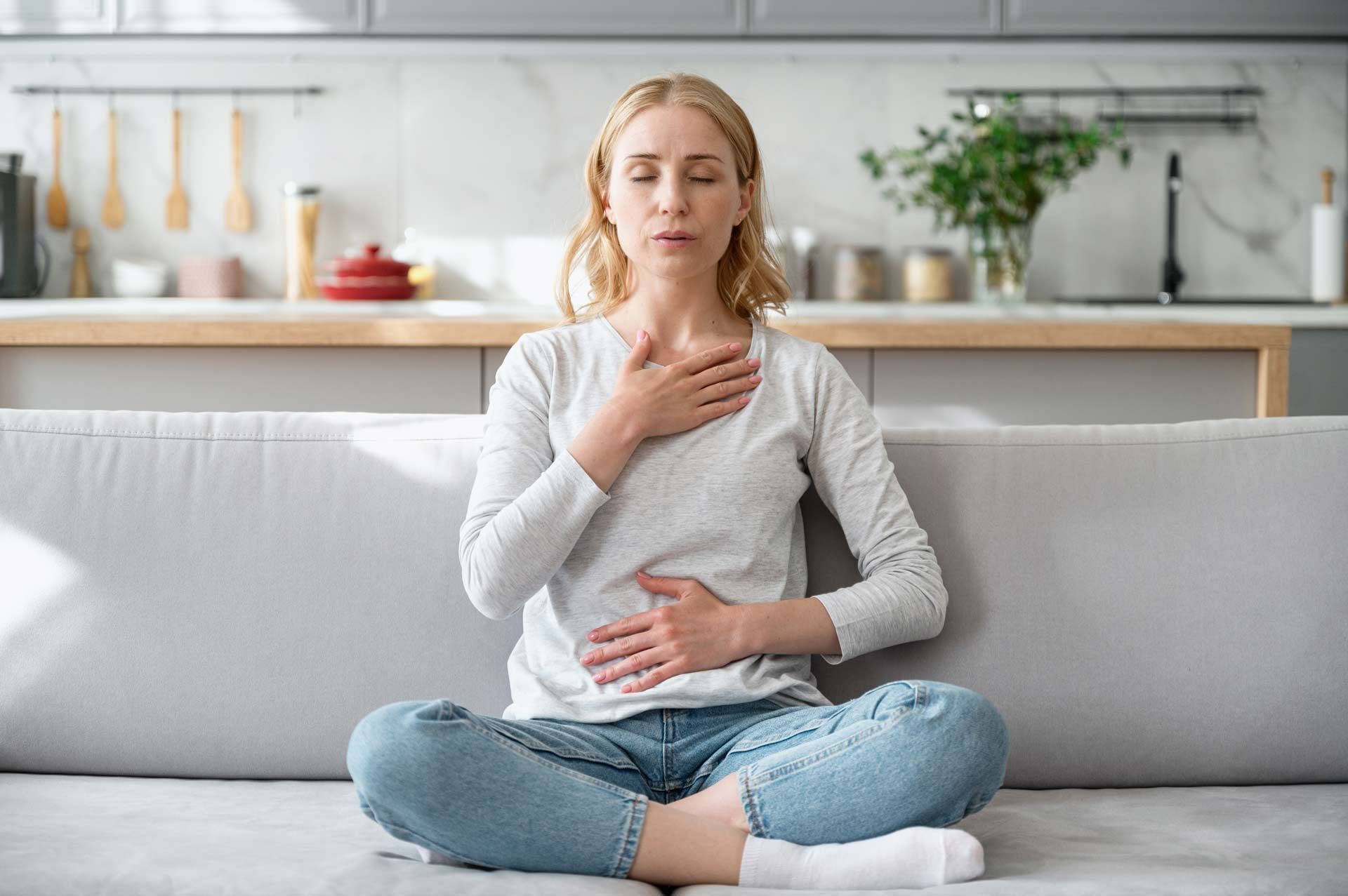Woman sitting on a couch with eyes closed, hands on chest and stomach, practicing breathing exercises.