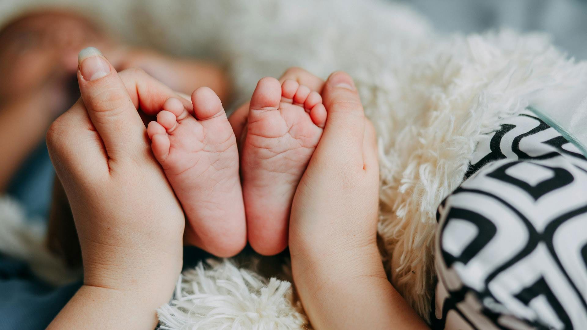 Hands gently cradle two tiny baby feet on soft, white blanket.