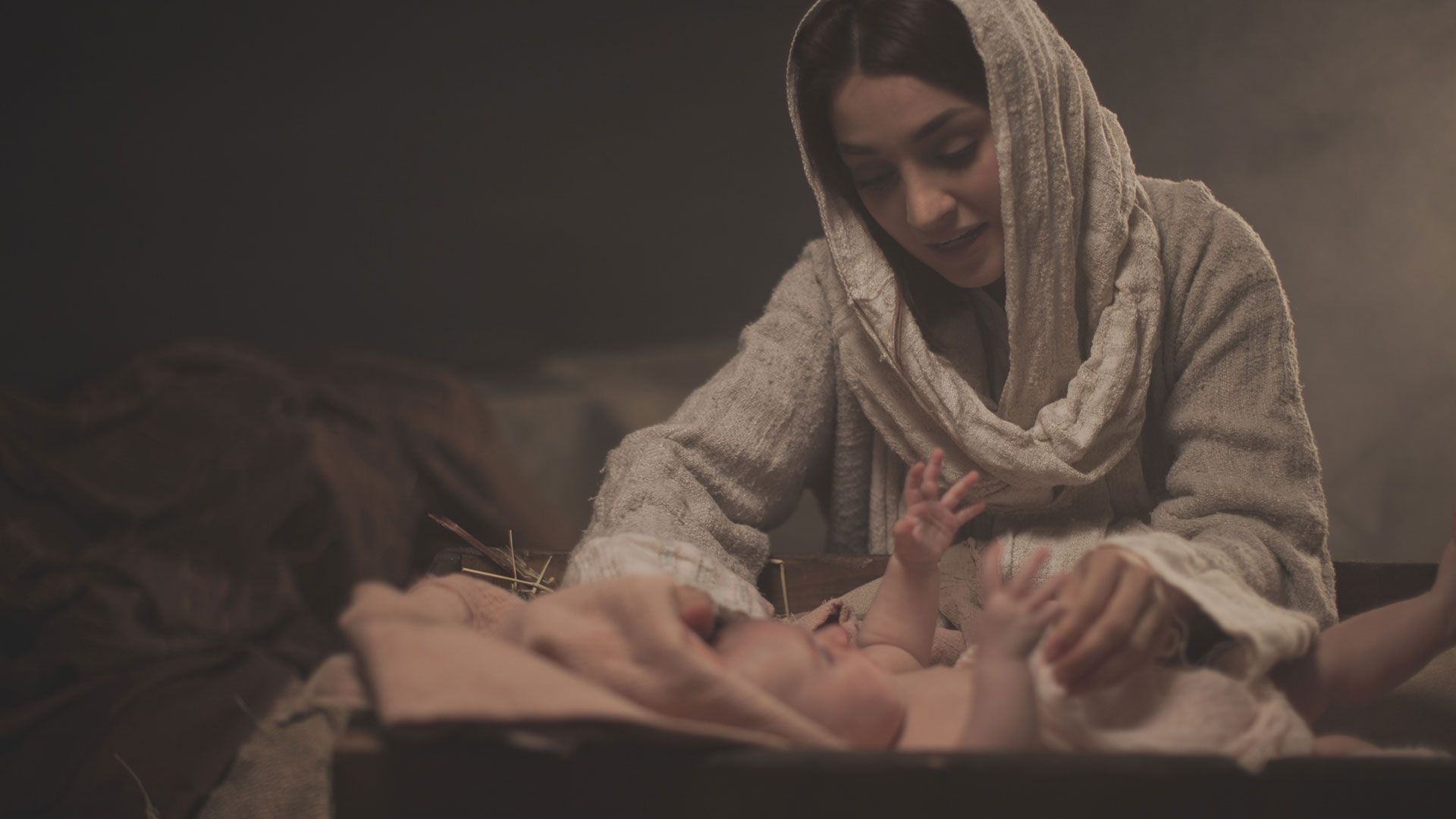 Woman in headscarf with baby in a manger, hands touching baby; dim lighting.