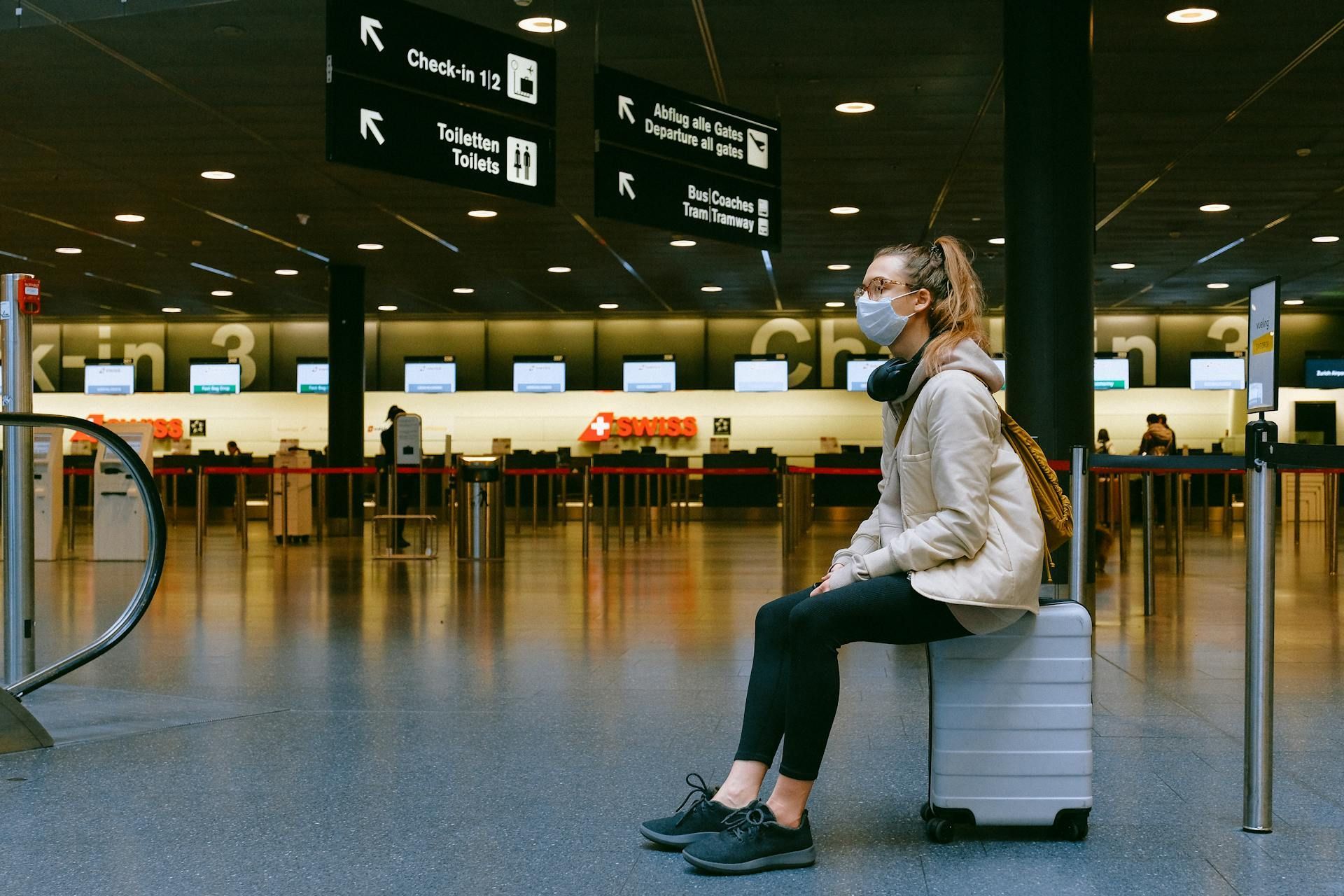 Woman in mask sits on suitcase in an empty airport. Black pants, cream jacket, headphones, check-in desks behind.