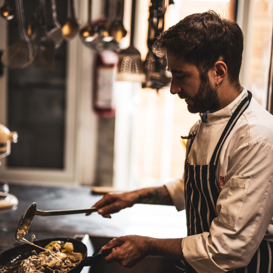 Cuoco in camice bianco e grembiule a righe, mentre mescola il cibo in una padella con un mestolo in cucina.