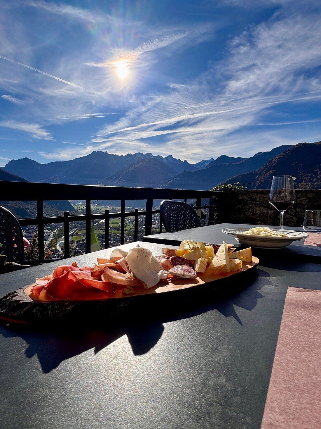 Tagliere di salumi su un tavolo con vista sulle montagne, sole splendente e cielo azzurro.