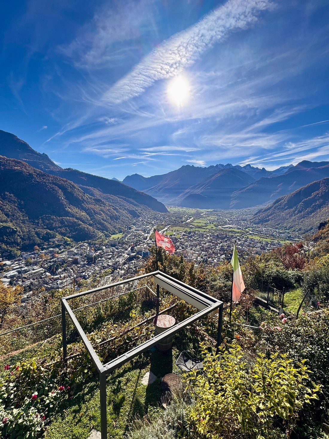 Vista panoramica di una cittadina incastonata tra le montagne, sotto un sole splendente e un cielo azzurro. Bandiere italiane.