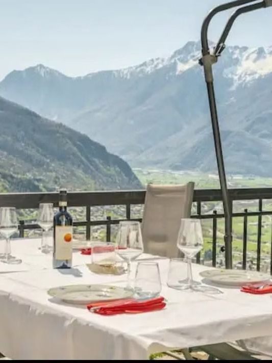 Tavolo apparecchiato per un pasto su un balcone con vista su una valle di montagna, bottiglia di vino e bicchieri in vista.