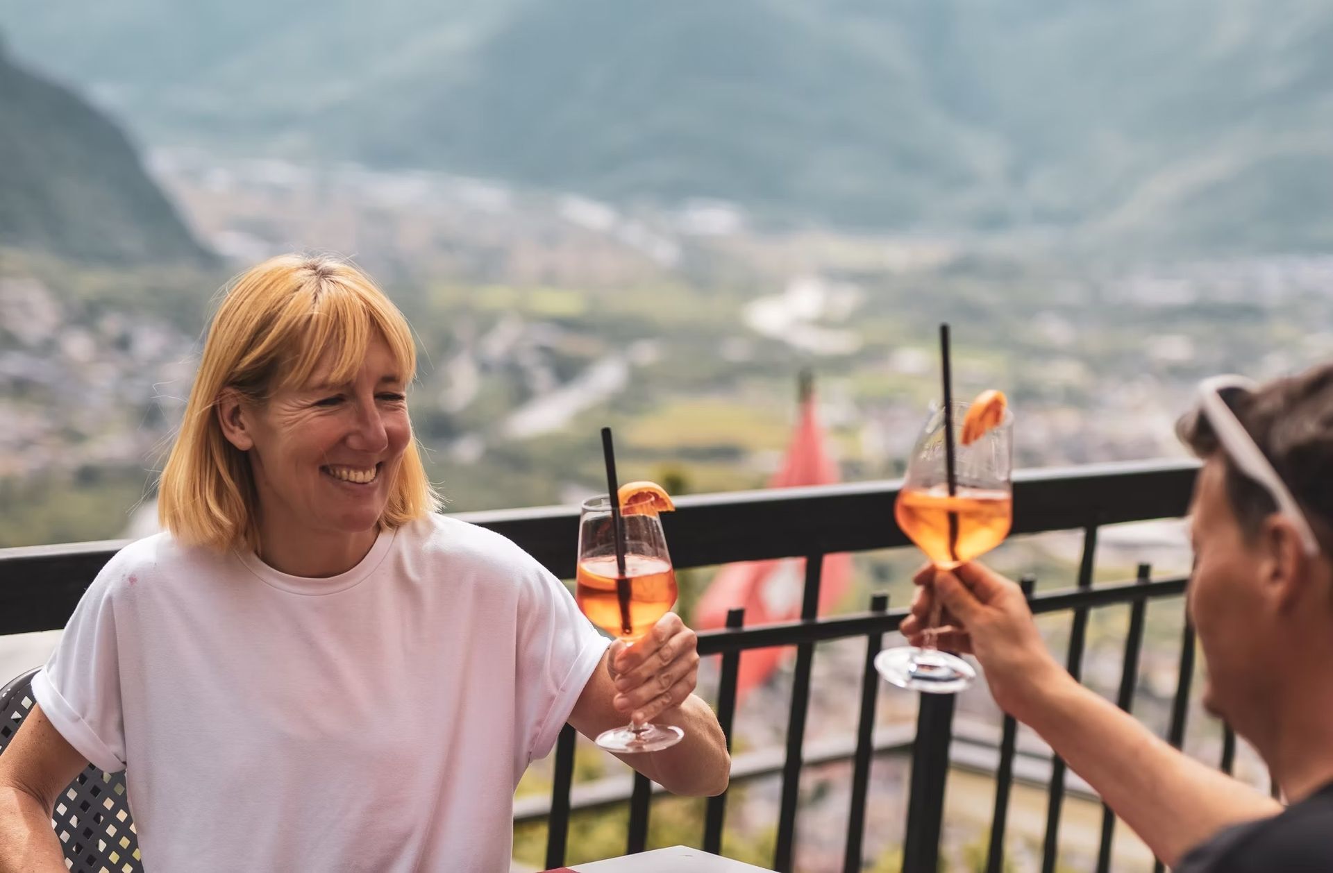 Donna sorridente che brinda con un drink su un balcone con vista su un paesaggio montuoso.