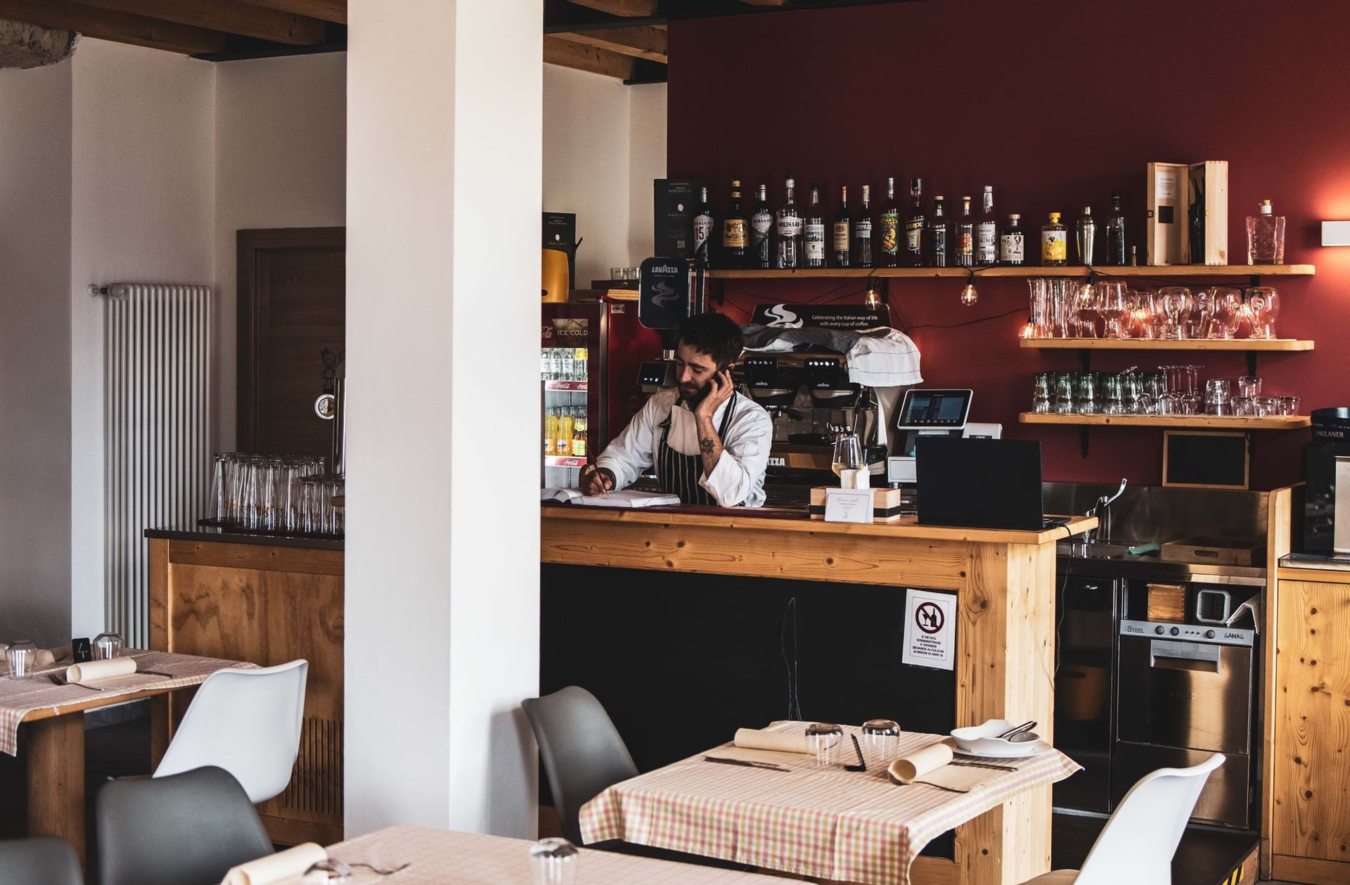 Interno di un ristorante con un barista al bancone che prende un ordine. Tavoli e sedie in primo piano.