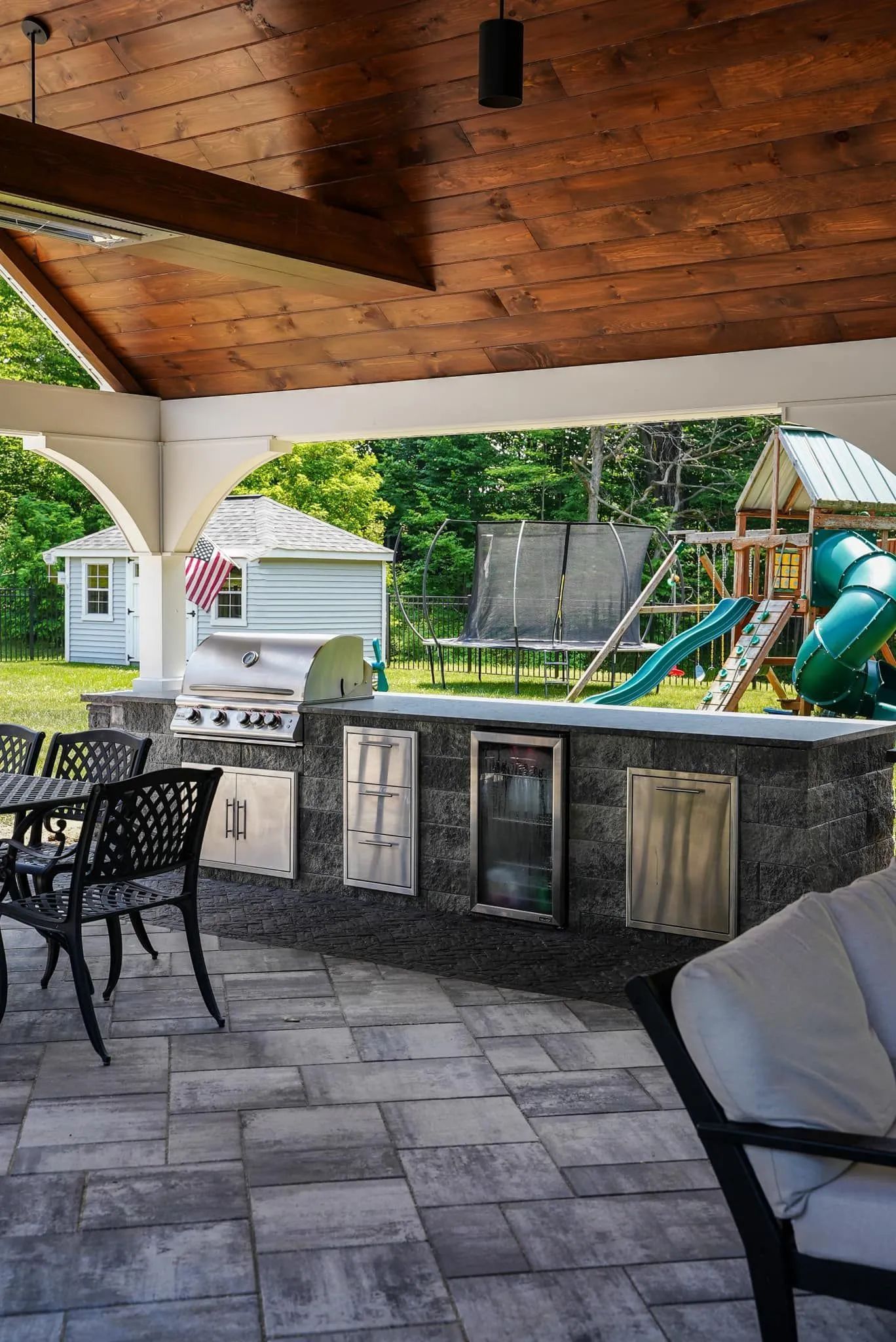 Patio with outdoor kitchen, dining set, and play area under a wooden roof.