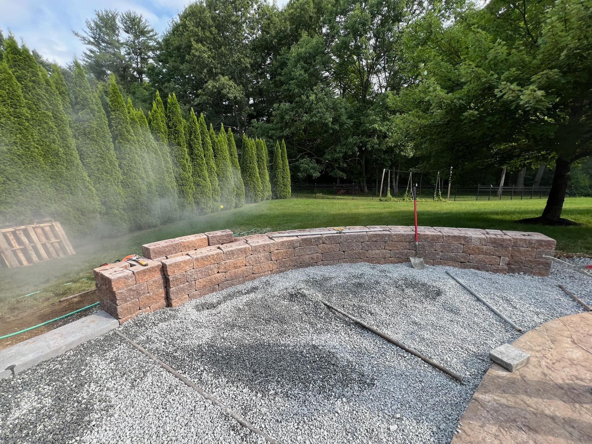 A brick wall is being built in a backyard with gravel and trees in the background.