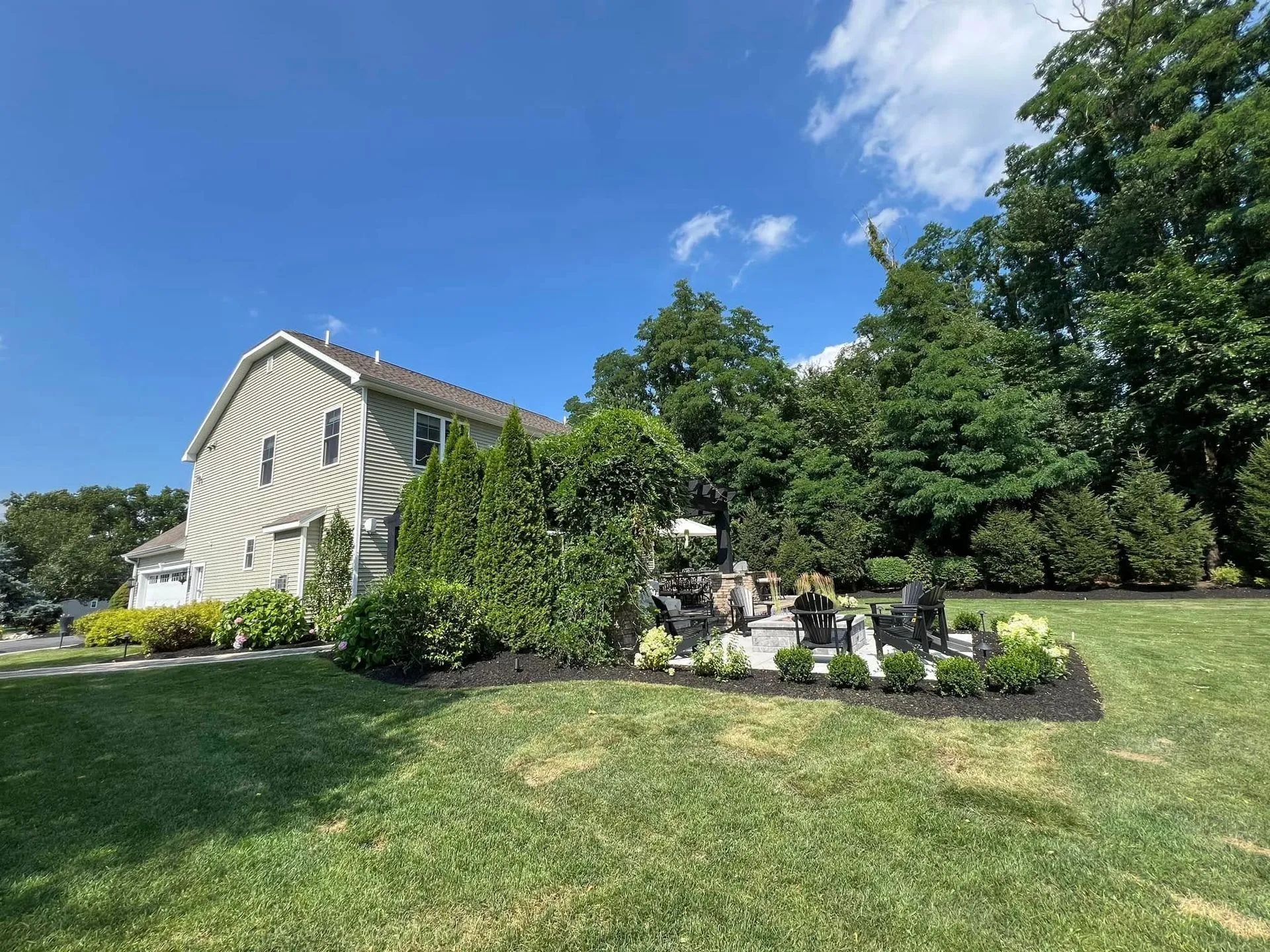 House with lawn and garden, a small patio, and trees under a blue sky.