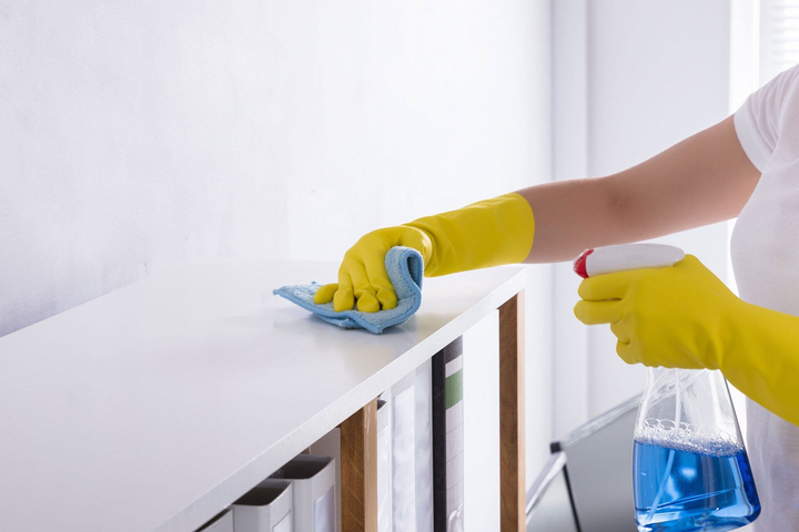 A woman wearing yellow gloves is cleaning a bookshelf with a cloth and spray bottle.