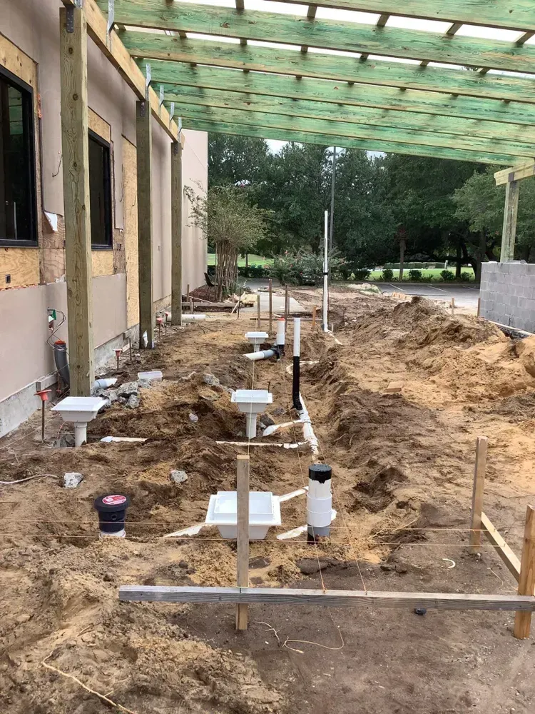 Construction site with exposed plumbing in a trench next to a building. Wooden pergola overhead.