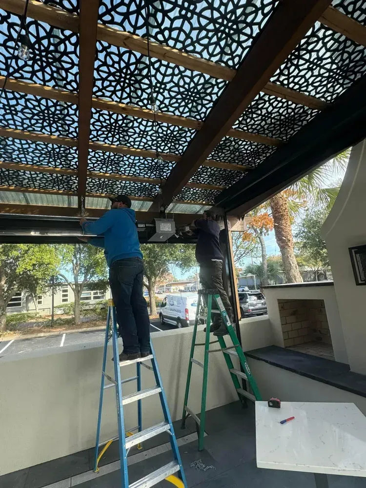 Two people on ladders install fixtures under a decorative wooden pergola.