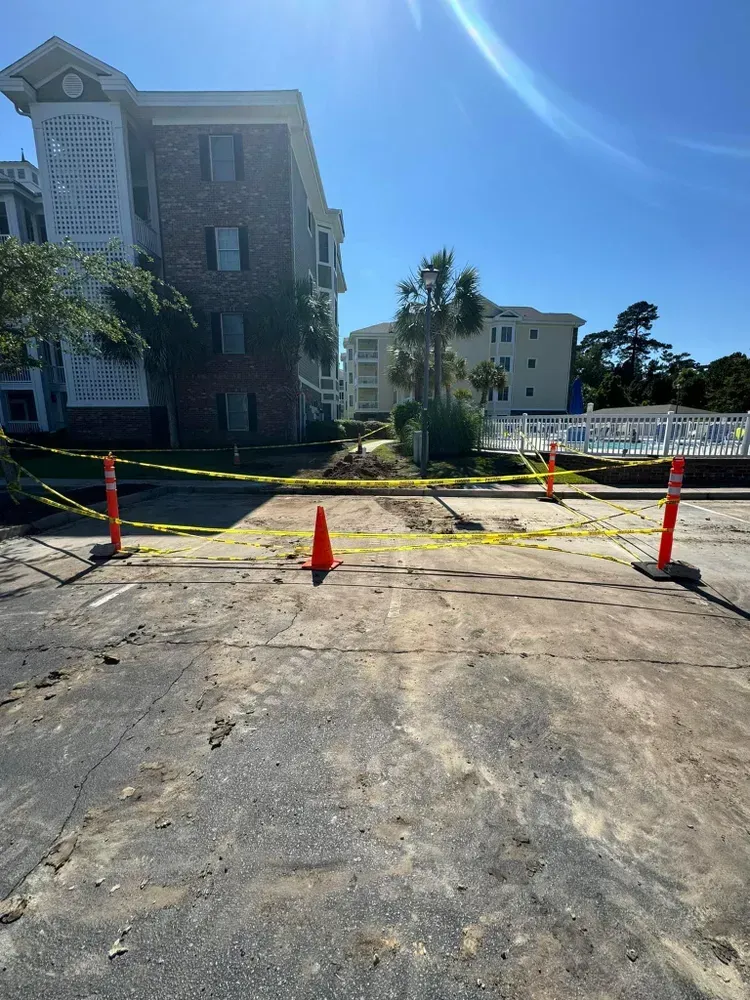 Road construction site with caution tape, cones, and buildings in the background. Sunny day.