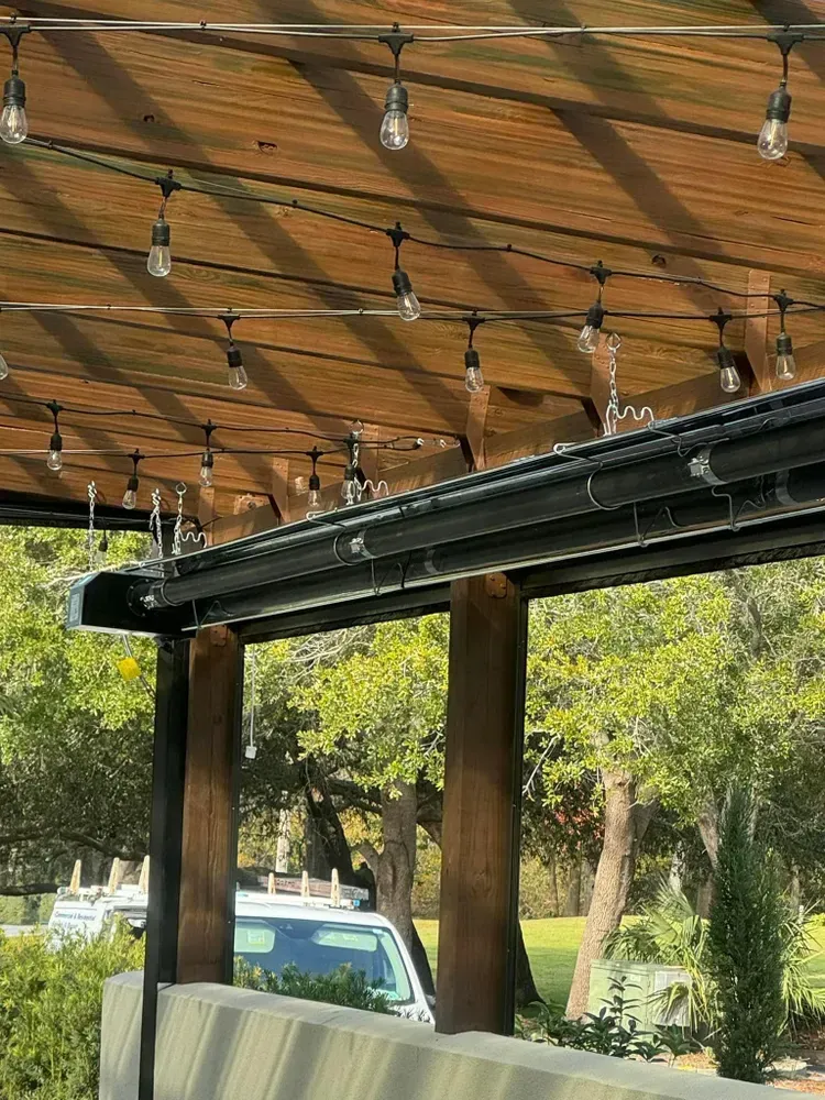 Pergola with string lights over a seating area, with trees and a vehicle in the background.